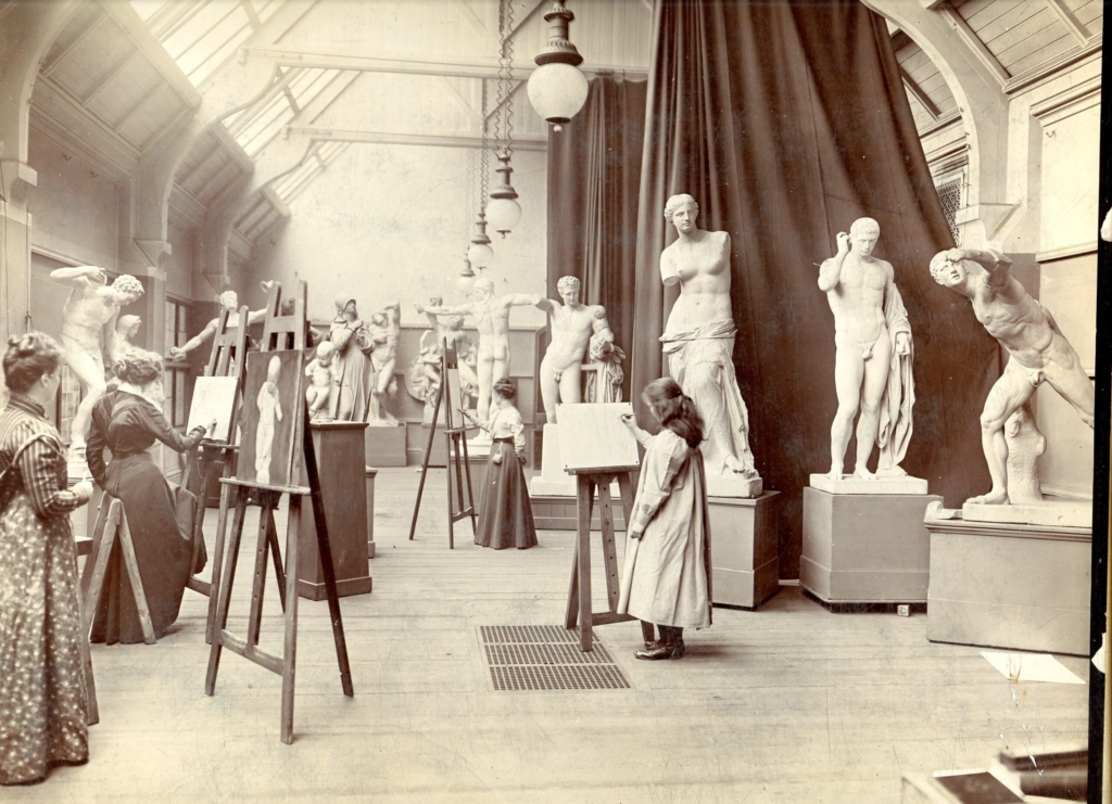 A vintage black-and-white photo of women and girls sketching classical nude statues in an art studio with high ceilings and skylights. They stand at easels, focused on their drawings