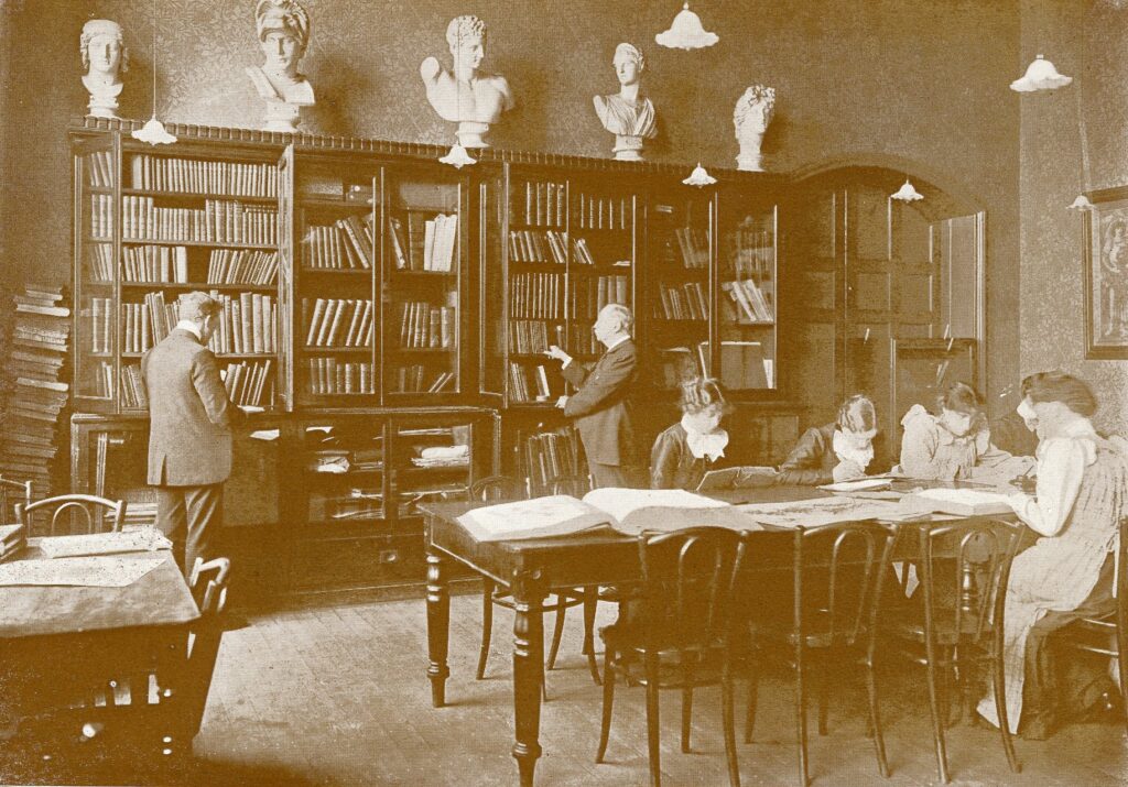 A sepia-toned image of a library with men browsing bookshelves and women seated at tables reading large open books, surrounded by tall shelves and classical busts above them.